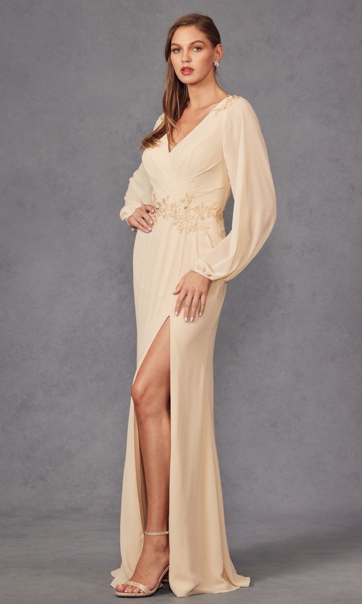 A woman wears the Juliet JTM16A V-Neck Long Sleeve Prom Gown in light beige, showcasing a gold embroidered waist and thigh-high slit, paired with nude heels against a gray backdrop.
